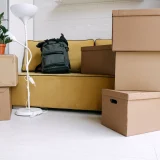 Cartons de déménagement empilés dans un salon avec canapé, lampadaire et sac à dos