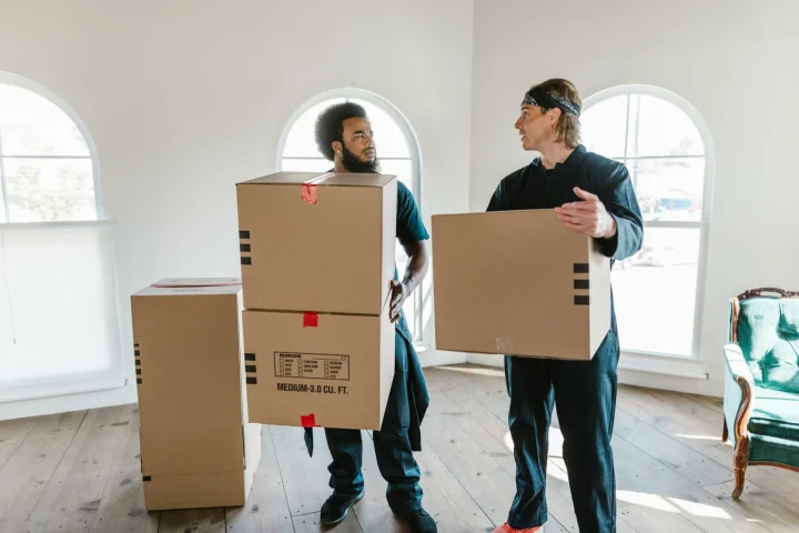 Deux déménageurs en combinaison portent des cartons dans un logement lumineux aux grandes fenêtres cintrées